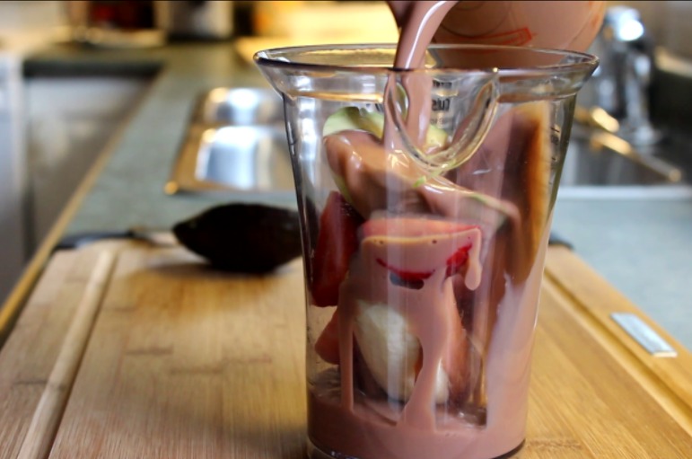 Pouring Chocolate Milk over Avocado, strawberries, and banana in a blender cup.
